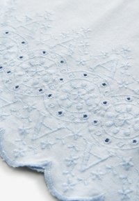 Světlomodrá látka s propracovanou výšivkou zobrazující kruhové a květinové vzory, zvlněný okraj a malé děrování.