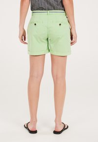 Lichtgroene shorts met een opgerolde zoom, voorzien van achterzakken met knoopsluitingen. Draagt zwarte slippers.