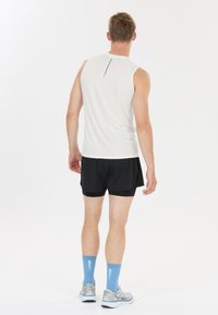 Man standing facing away wearing a white sleeveless shirt, black shorts, light blue socks, and gray running shoes on a white background.
