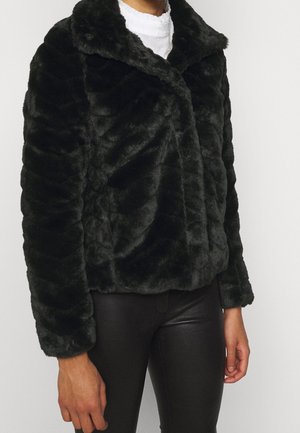 Person trägt eine schwarze Kunstpelzjacke mit weitem Kragen über einem weißen Hemd und schwarzen Lederhosen vor einem weißen, schlichten Hintergrund.