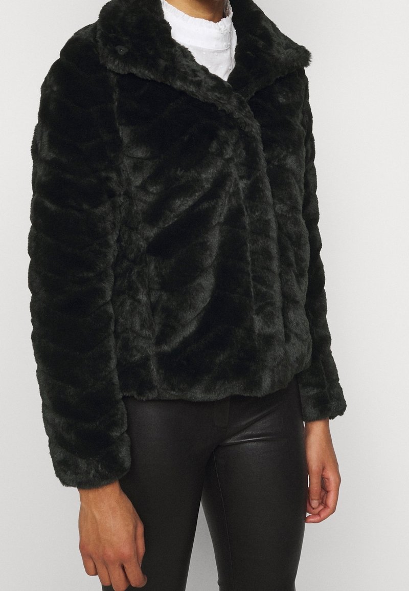 Personne portant une veste en fausse fourrure noire à large col sur une chemise blanche et un pantalon en cuir noir sur un fond blanc uni.