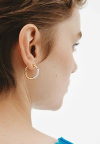 Pendiente de aro dorado con un acento blanco. Forma circular, textura suave. Lleva puesto un modelo de cabello corto, mostrando el diseño de la oreja y el pendiente.