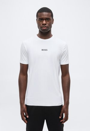 Bărbat purtând un tricou alb cu mâneci scurte, cu inscripția „BOSS” scrisă cu negru pe piept, stând în fața unui fundal clar, simplu.