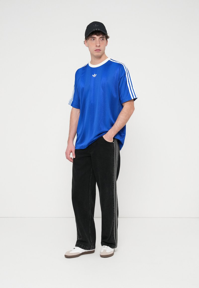 Blaues Sport-T-Shirt mit weißem Kragen und drei weißen Streifen an den Ärmeln, kombiniert mit schwarzen Hosen mit Seitenstreifen und weißen Sneakers.