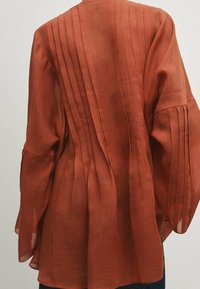Rustfarvet linnedbluse med lange, brede ærmer og vertikale plisseringer langs ryggen, med en let løs silhuet.