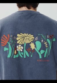 Personne portant une chemise bleu foncé avec un motif floral stylisé coloré et le texte « BOBO CHOSES » dans le dos.