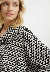 Zwarte en crèmekleurige blouse met een geometrisch patroon van in elkaar verweven vormen. Gemaakt van lichtgewicht stof met een losse, gedrapeerde kraagontwerp.