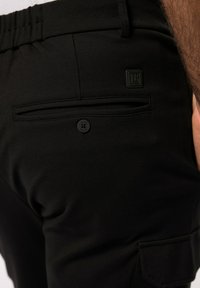 Schwarze Stretchhose mit glatter Textur, ausgestattet mit einer Gesäßtasche und einem Knopfverschluss, sowie einem kleinen Logostreifen in der oberen Ecke.