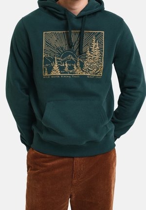 Personne portant un sweat à capuche vert foncé avec un graphique jaune de sentier de randonnée montrant des arbres et des montagnes, associé à un pantalon en velours côtelé marron.