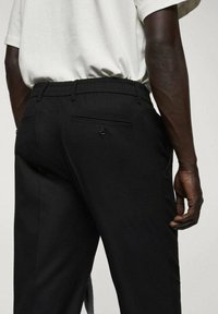 Homme portant un pantalon noir sur mesure avec une poche arrière boutonnée et une chemise blanche à manches courtes rentrée, debout avec la main droite détendue.