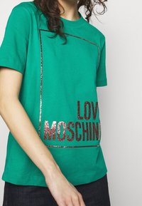 Person som bär en grön kortärmad skjorta med glittrande röd text "LOVE MOSCHINO" delvis synlig innanför en tunn rektangulär kontur.