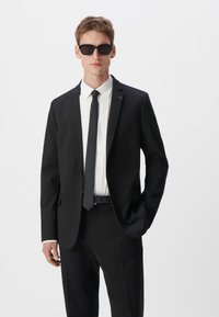 Czarny garnitur z białą koszulą, czarnym krawatem i okularami przeciwsłonecznymi. Slim fit, gładki materiał i subtelny połysk tworzą formalny wygląd.