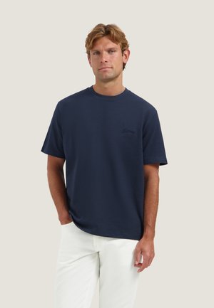 Mann mit hellbraunen Haaren, der ein marineblaues kurzärmeliges Hemd und weiße Hose trägt, steht vor einem einfachen hellen Hintergrund.