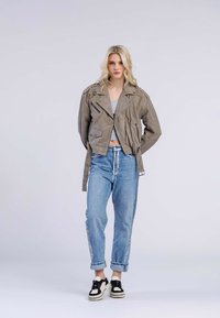 Grijze suède motorjack met ritsen en schouderepauletten, gecombineerd met lichtblauwe high-waisted jeans en zwarte sneakers.