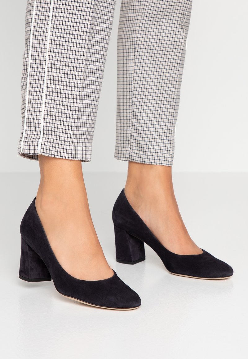 Femme portant des chaussures en daim noir à talons carrés et un pantalon blanc coupé avec un petit motif à carreaux et des bandes latérales.