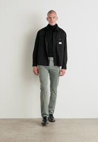Jachetă neagră peste un pulover negru cu guler înalt, asortată cu pantaloni gri deschis. Modelul poartă pantofi negri, stând pe un fundal neutru.
