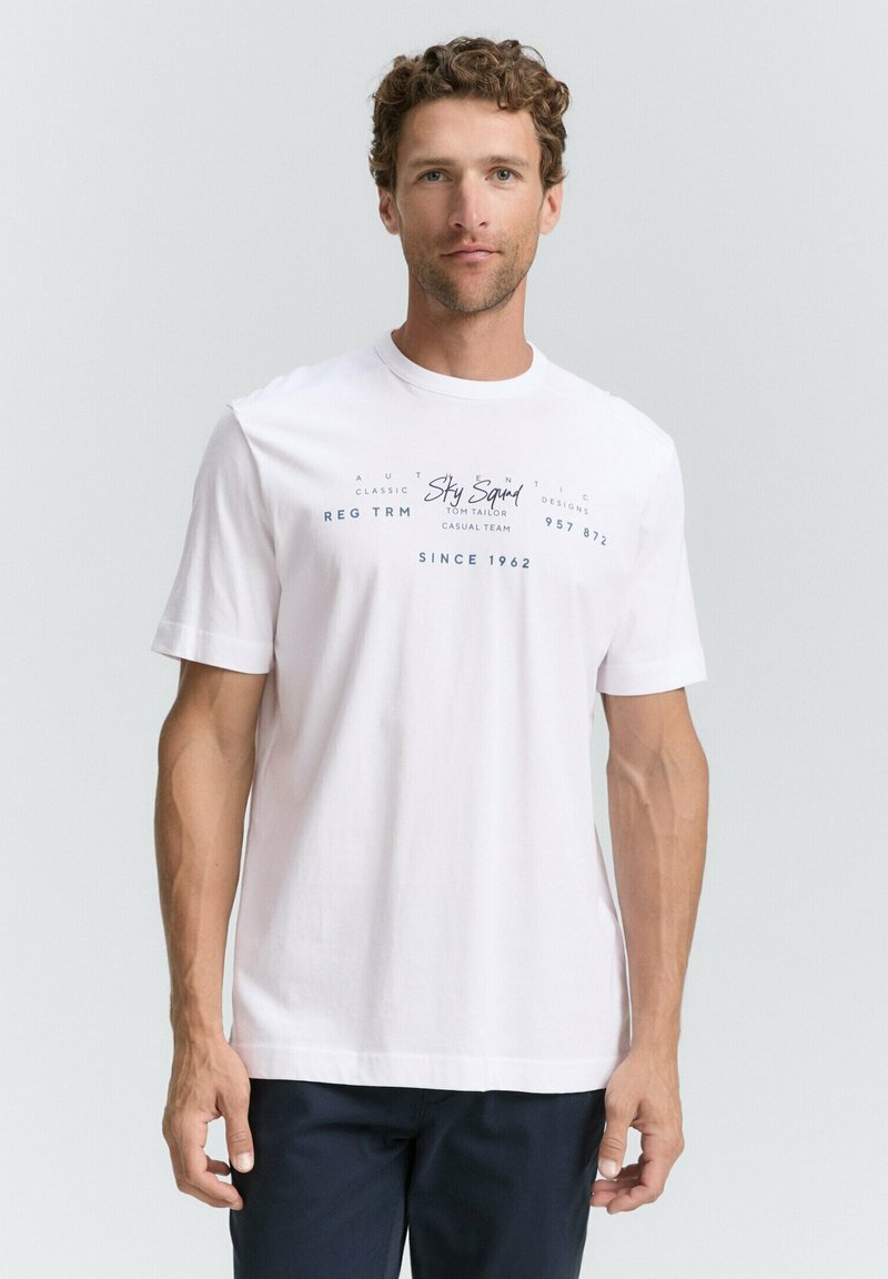 Hvid bomulds-T-shirt med korte ærmer, rund halsudskæring og blå tekstgrafik på forsiden. Båret med mørke bukser.