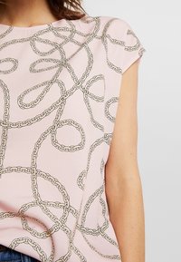 Light pink short-sleeve top featuring a chain pattern outlined in dark tones. Fabric appears smooth with a relaxed fit.