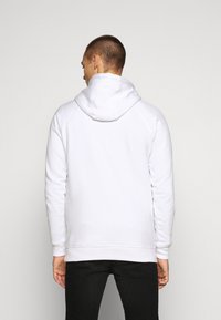 Balta pūkinė su gobtuvu su kengūros kišene, tekstūruotas audinys, reguliuojamas raištelis ant gobtuvo ir raibos rankovės. Atgalinis vaizdas su juodais džinsais.