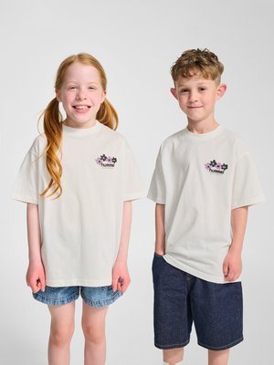 Uśmiechnięta dziewczyna z rudymi włosami w jeansowych szortach i chłopiec z kręconymi włosami w ciemnych szortach, oboje ubrani w białe koszulki z kwiatowym logo, stojący obok siebie.