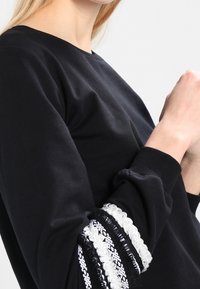 Svart sweatshirt med texturerade ärmar med silver- och vita dekorationer. Ribbad mudd och avslappnad passform. Slät tygfinish.