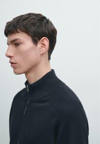 Schwarzer Reißverschluss-Pullover mit geripptem Kragen und Schulterakzenten, glatter Textur und minimalistischem Design. Wird von einem seitlich ausgerichteten Modell getragen.