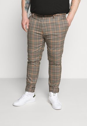 Mann trägt beige, braune und schwarze karierte Hose, weiße Sneaker und ein schwarzes Hemd und steht mit den Händen in den Taschen.