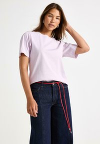 Lyse lilla bomulds T-shirt med rund hals og korte ærmer, parret med mørk denim brede bukser og en rød snor.