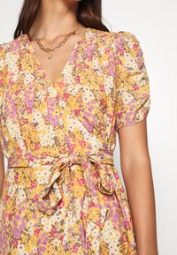 Femme portant une robe portefeuille à fleurs jaunes avec des manches bouffantes et une ceinture à nœud, accessoirisée avec des colliers en chaîne dorée superposés.