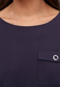 Marineblaues Kleidungsstück mit einem runden Ausschnitt und einer Brusttasche mit einem silbernen Ösen-Detail, aus einem glatten, leichten Stoff hergestellt.