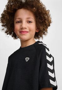 T-shirt nera con colletto rotondo, caratterizzata da strisce a zigzag bianche sulle maniche e da un piccolo logo sul petto. Texture di cotone liscia.