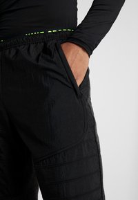 Schwarze Sporthose mit strukturiertem Finish, seitlichen Taschen und einem grünen Akzent am Bund. Das Material wirkt leicht und strapazierfähig.