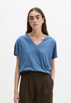 Jeune femme aux longs cheveux bruns portant un t-shirt bleu ample à col en V et un pantalon marron foncé taille haute, debout devant un fond blanc uni.