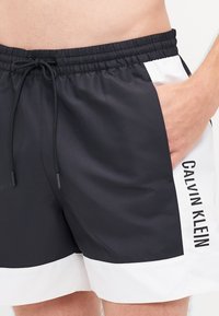 Črnovbele kopalke z elastičnim pasom, vrvico, stranskima žepoma in odtisnjenim napisom "CALVIN KLEIN" s strani.