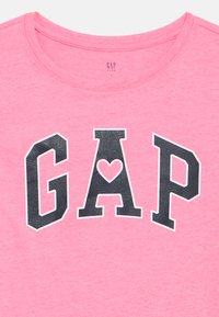 Rózsaszín gyerekpóló fekete, nagy „GAP” betűkkel, az "A" betű belsejében egy szív alak, kerek nyakkivágás alatt.