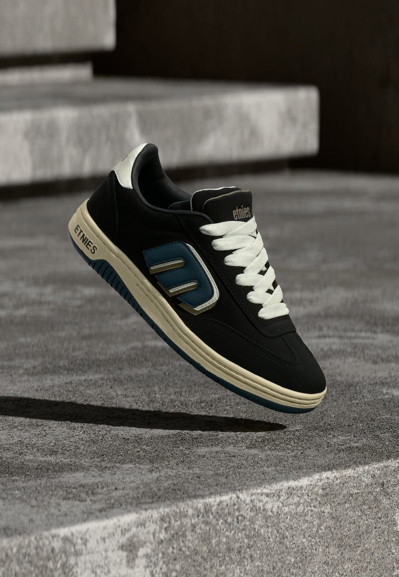 Μαύρο αθλητικό παπούτσι με ανοιχτό γκρι σόλα και μπλε λεπτομέρειες. Διαθέτει άσπρα κορδόνια, στρογγυλή σχήμα και λογότυπο στο πλάι και στην γλώσσα.