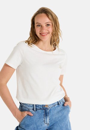 T-shirt en coton blanc à manches courtes, col rond et détail brodé doré sur la poitrine. Associé à un jean taille haute bleu clair.