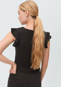 Top nero con maniche a volant, tessuto texture, silhouette aderente e scollatura rotonda. Capelli lunghi raccolti in una coda di cavallo, con orecchini a cerchio.
