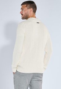Cremfarbener gerippter Pullover mit langen Ärmeln und hohem Kragen. Mit einem kleinen schwarzen Etikett auf der Rückseite. Kombiniert mit grauen Hosen.