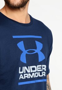 Granatowa bawełniana koszulka z dużym niebieskim logo Under Armour i białym napisem. Klasyczny krój z okrągłym dekoltem, krótki rękaw, gładka tekstura.