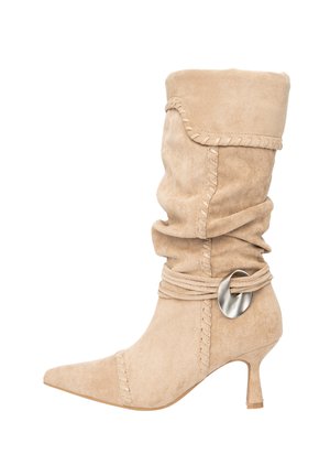 Stivaletto beige in camoscio con design morbido, punta appuntita, cuciture decorative e un dettaglio metallico su una cintura avvolta vicino alla parte superiore.