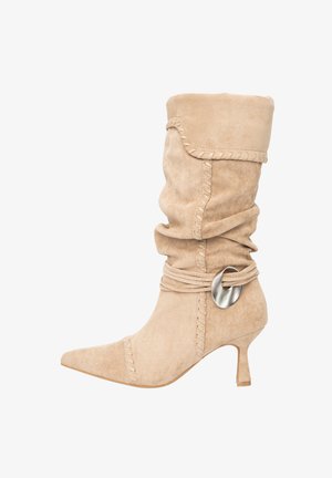 Bottine beige en daim avec un design souple, un bout pointu, des coutures décoratives et un accent métallique sur une ceinture enveloppante près du haut.
