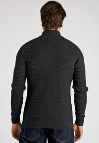 Schwarzer Strickpullover mit strukturiertem Muster, hohem Kragen und gerippten Bündchen sowie Saum. Gekleidet zu blauen Denim-Jeans.