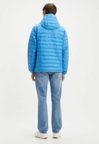 Doudoune bleu clair avec un col montant et une capuche zippée. Présente un motif matelassé horizontal. Portée avec un jean bleu clair et des chaussures blanches.