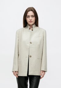 Γυναίκα με καστανά ίσια μαλλιά που φοράει ένα μπεζ σακάκι με ψηλό γιακά και κουμπιά, καθώς και μαύρο παντελόνι, στέκεται μπροστά σε λευκό απλό φόντο.