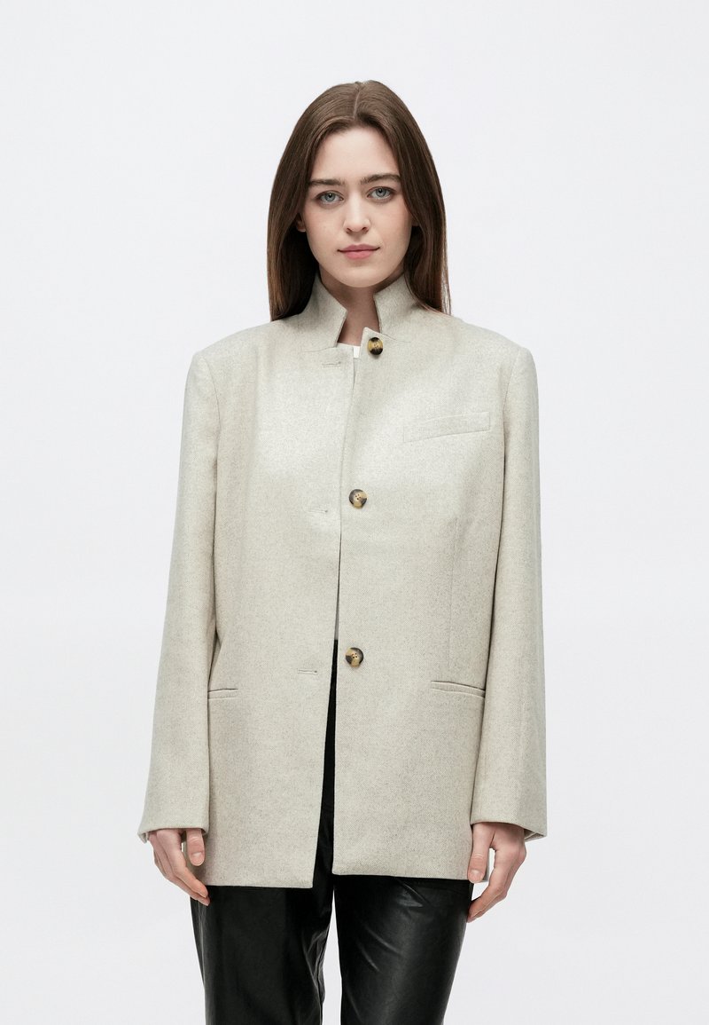 Γυναίκα με καστανά ίσια μαλλιά που φοράει ένα μπεζ σακάκι με ψηλό γιακά και κουμπιά, καθώς και μαύρο παντελόνι, στέκεται μπροστά σε λευκό απλό φόντο.