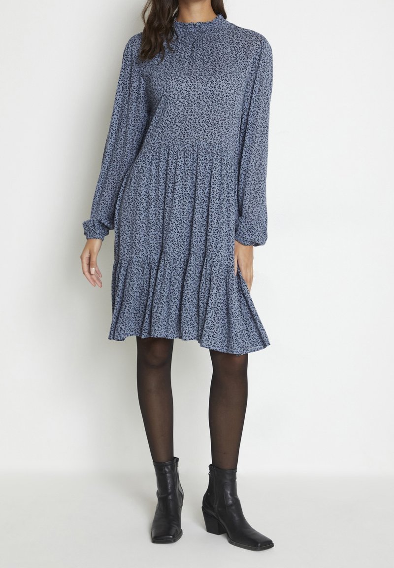 Mujer vestida con un vestido de manga larga hasta la rodilla de flores azules, con medias negras y botas negras hasta el tobillo, posando frente a un fondo blanco liso.