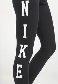 Svarta leggings av stretchigt tyg har stora vita bokstäver som stavar "NIKE" längs den yttre sömmen. Slät textur, åtsittande design.