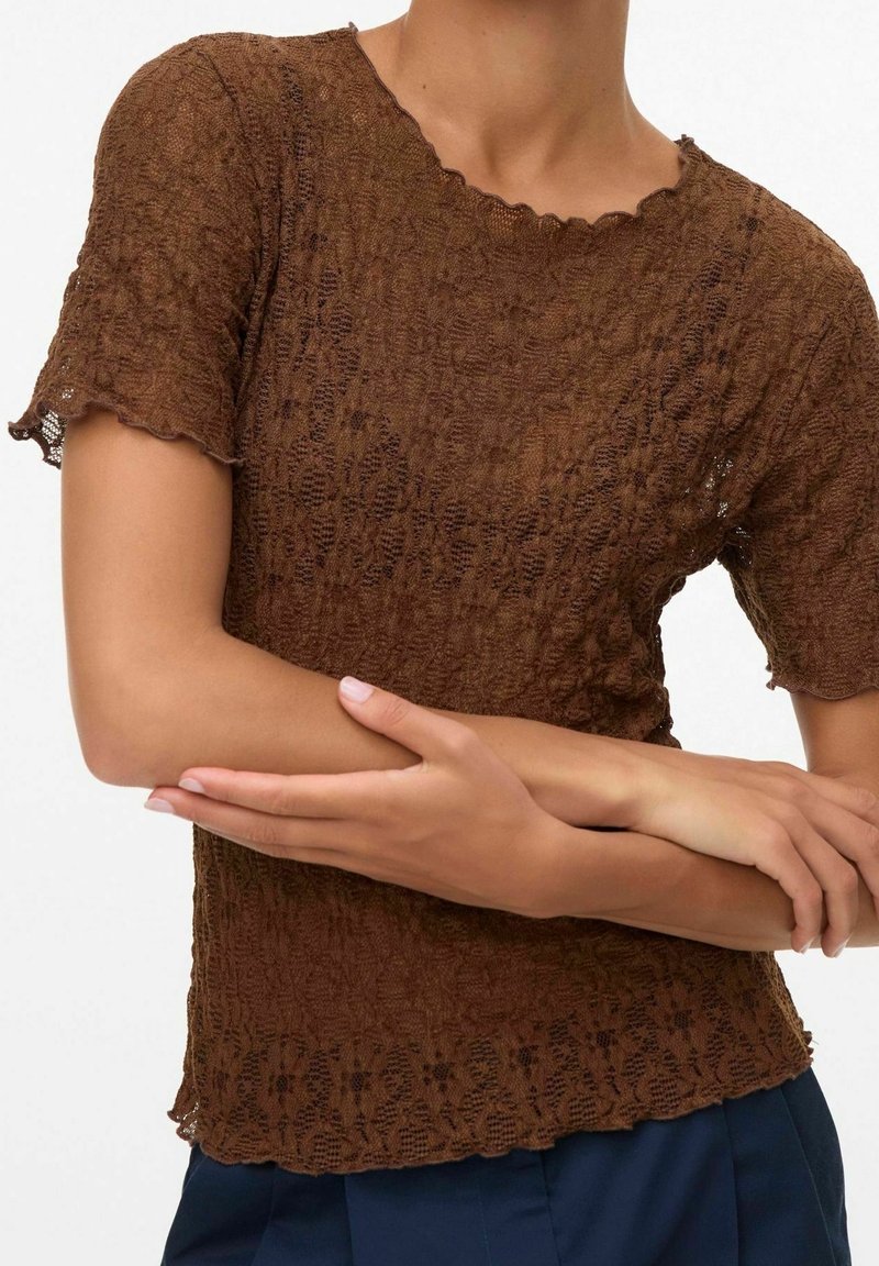 Femme portant un haut marron texturé à manches courtes avec des bords festonnés, les bras croisés, vue du cou aux hanches sur un fond uni.