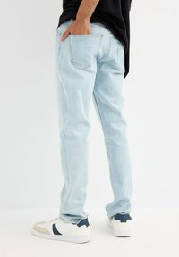 Jeans de mezclilla en azul claro con un corte relajado, que presentan bolsillos traseros clásicos y detalles de costura sutiles. Llevados con zapatillas blancas acentuadas con navy.